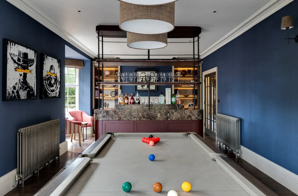 Pool table room with gray felt, racked red balls and colorful cue balls, navy walls, marble-topped home bar and pendant lights.