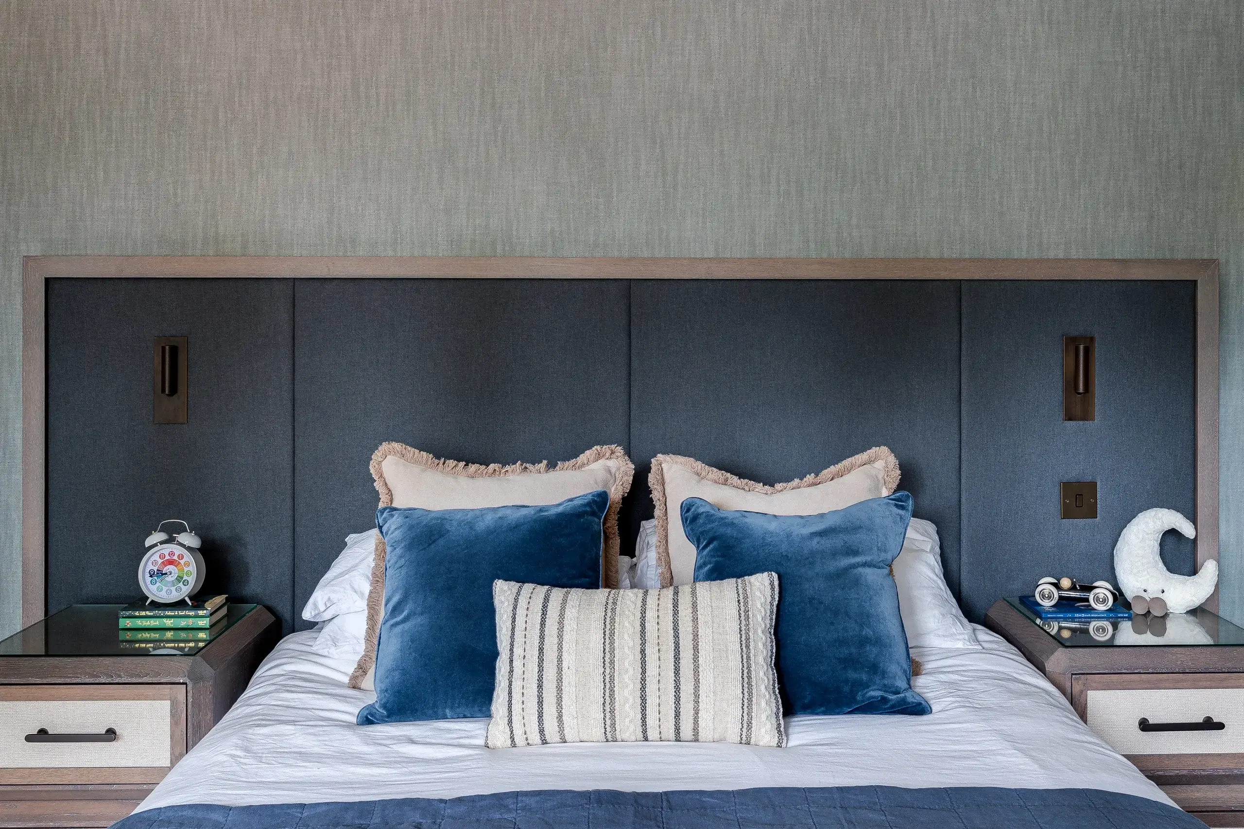 King bed with dark upholstered headboard, blue velvet throw pillows, striped lumbar pillow, white bedding and bedside tables with alarm clock and decorative items.