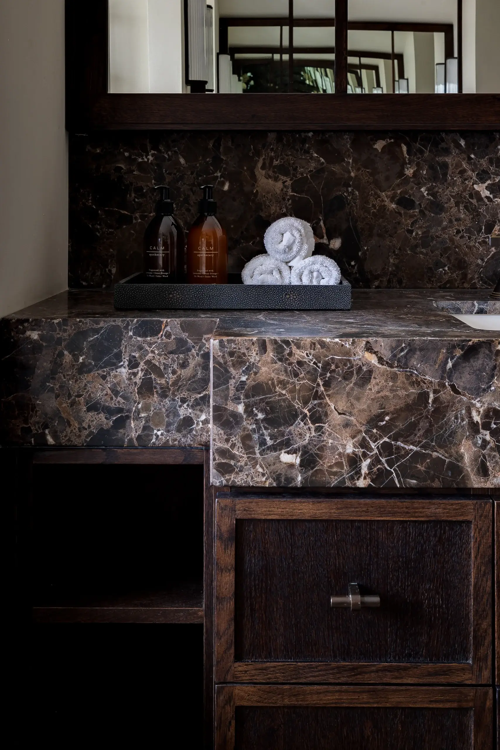 Dark brown marble bathroom vanity with rolled white towels and amber soap bottles on a black tray beneath a mirror