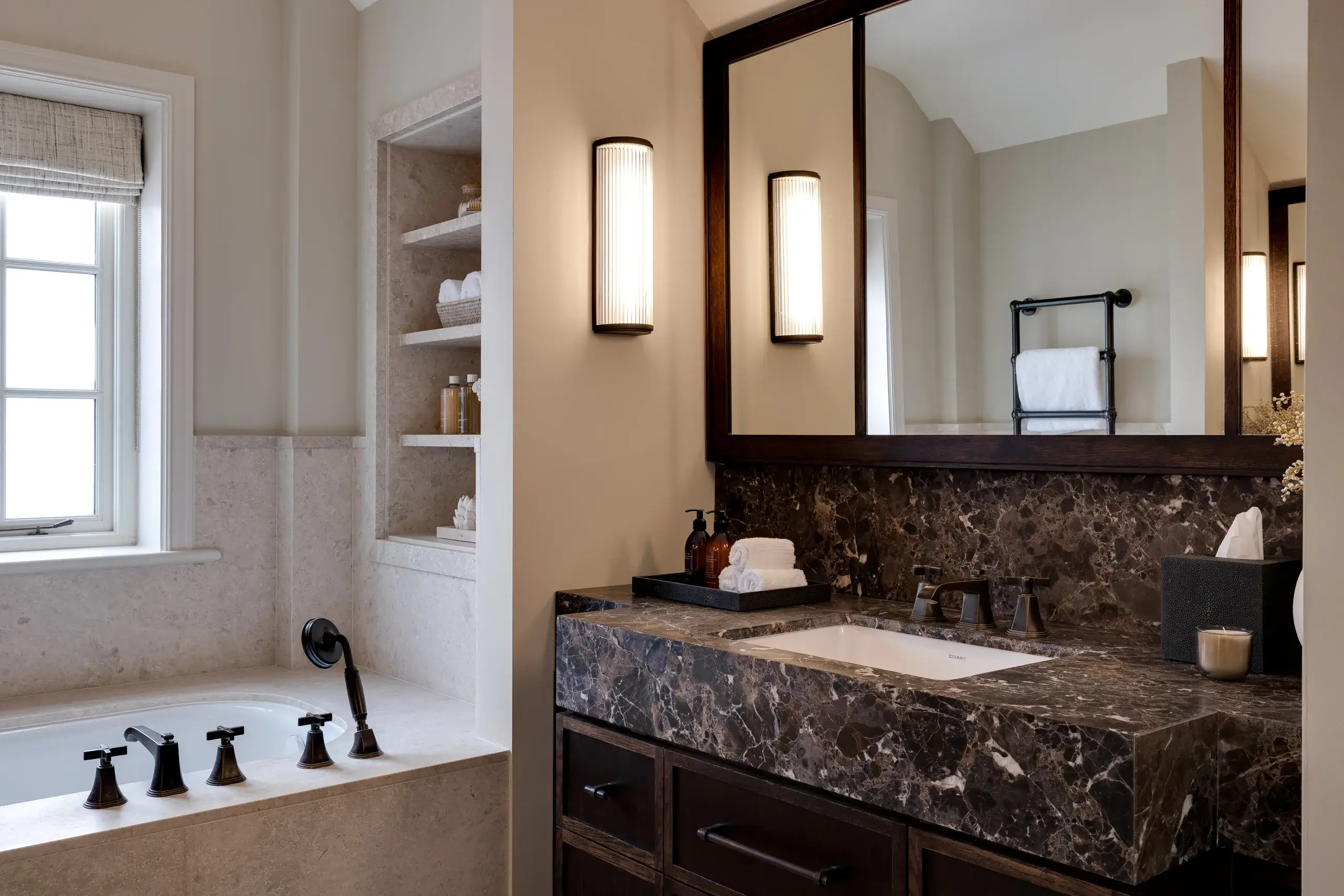 Luxurious bathroom with dark marble vanity, under-mount sink, wall mirror and sconce, beige tub with black fixtures and built-in shelving.