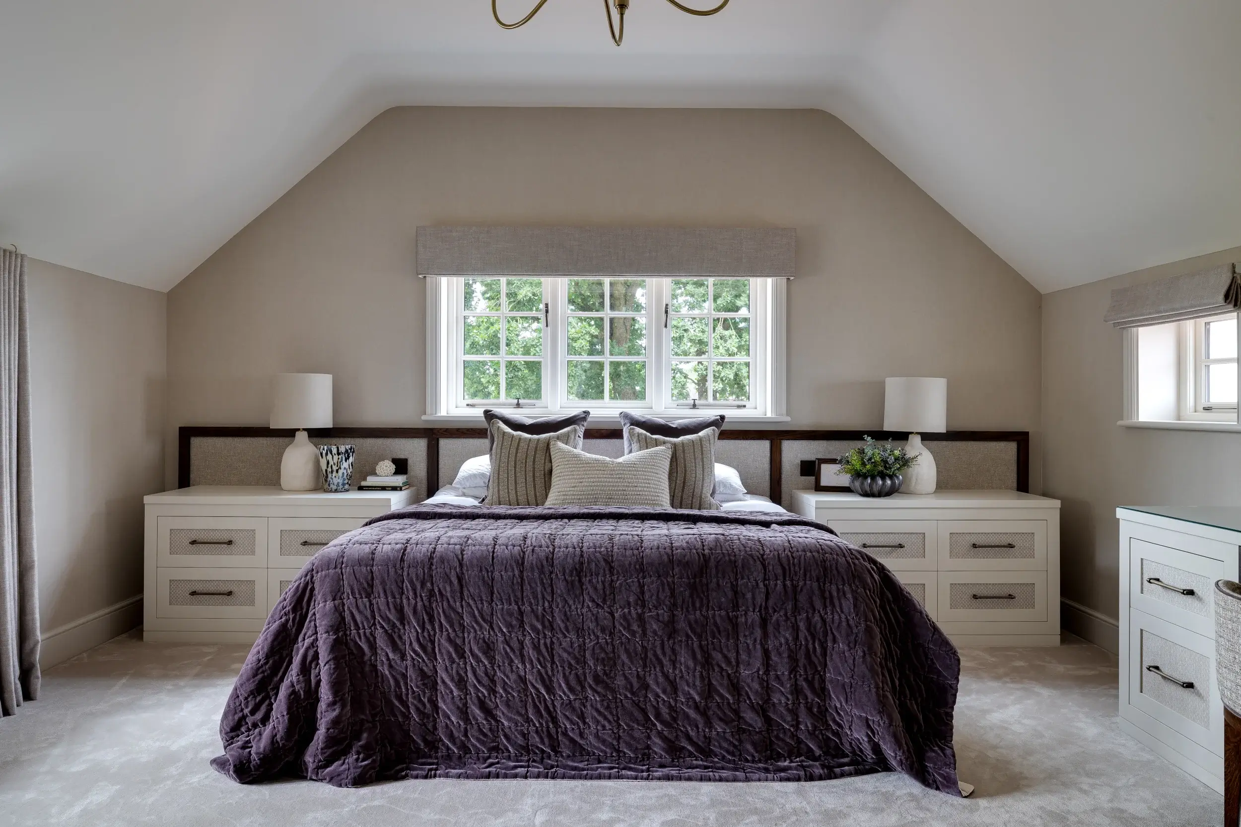Calming bedroom with pitched ceiling, soft neutral tones, and a deep plum bedspread framed by bespoke bedside cabinetry.