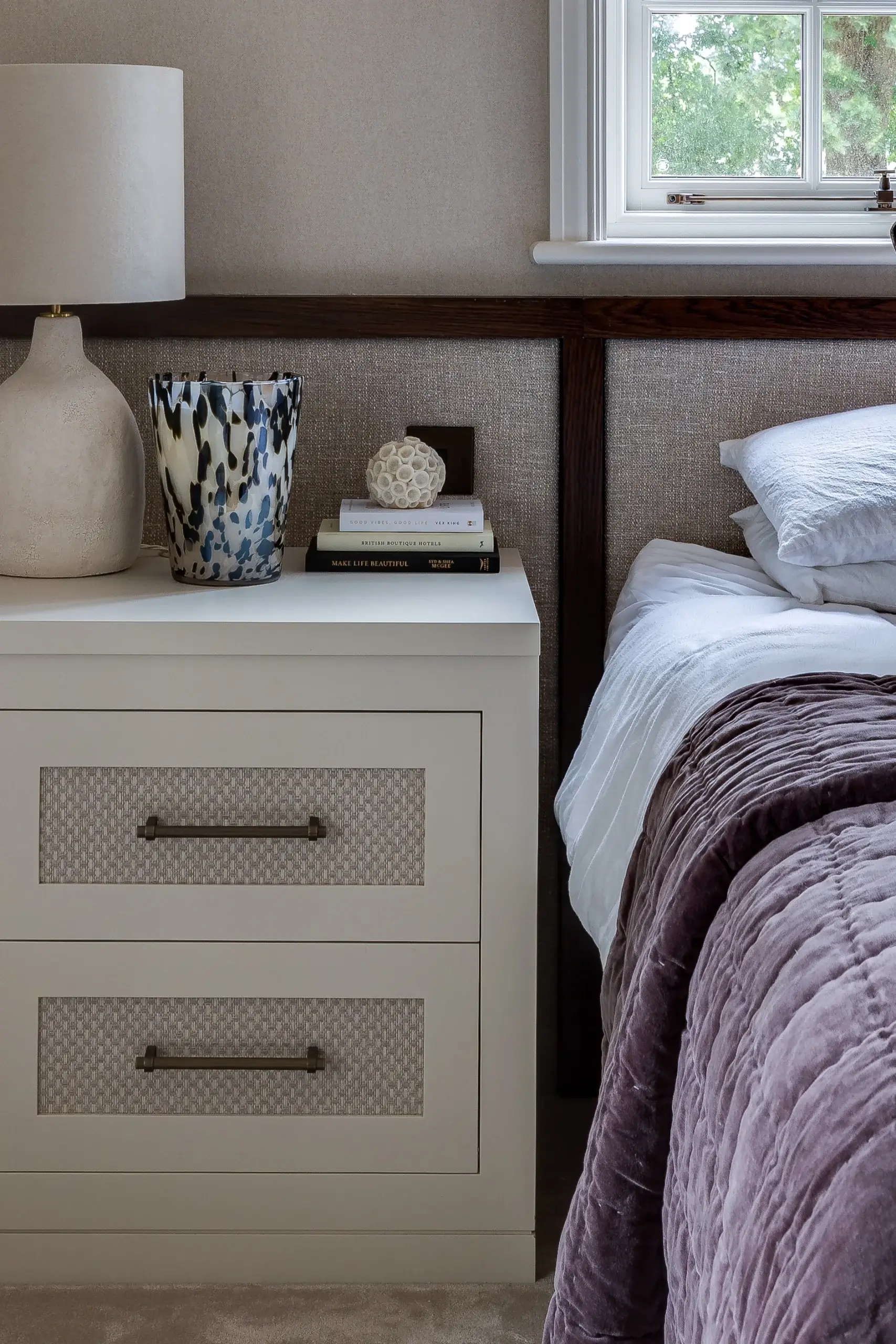 Bedside table with lamp, patterned vase, stacked books and decorative orb next to a bed with white sheets and mauve quilt.