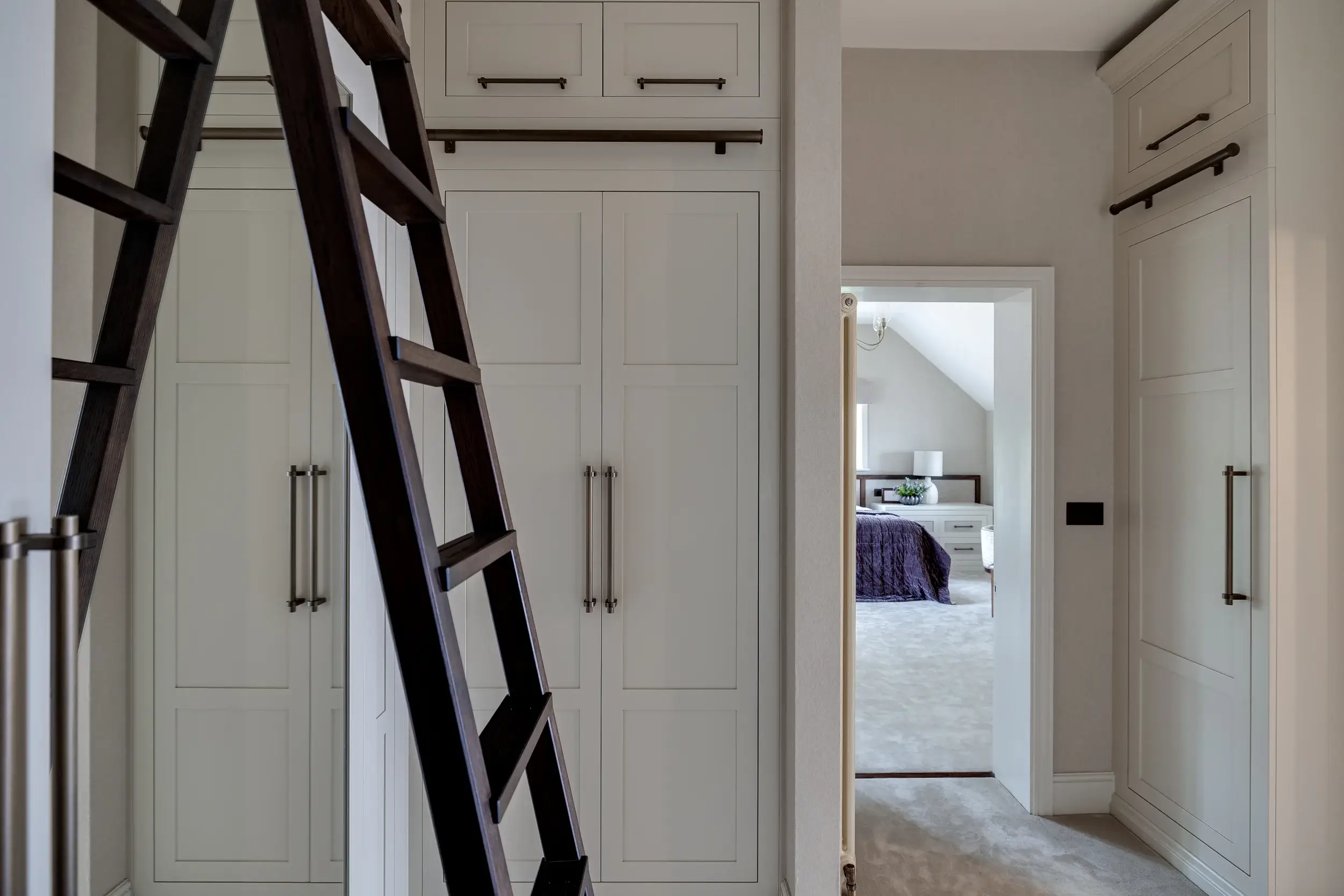 Bespoke wardrobe-lined hallway with sliding ladder, opening through to the master bedroom beyond.