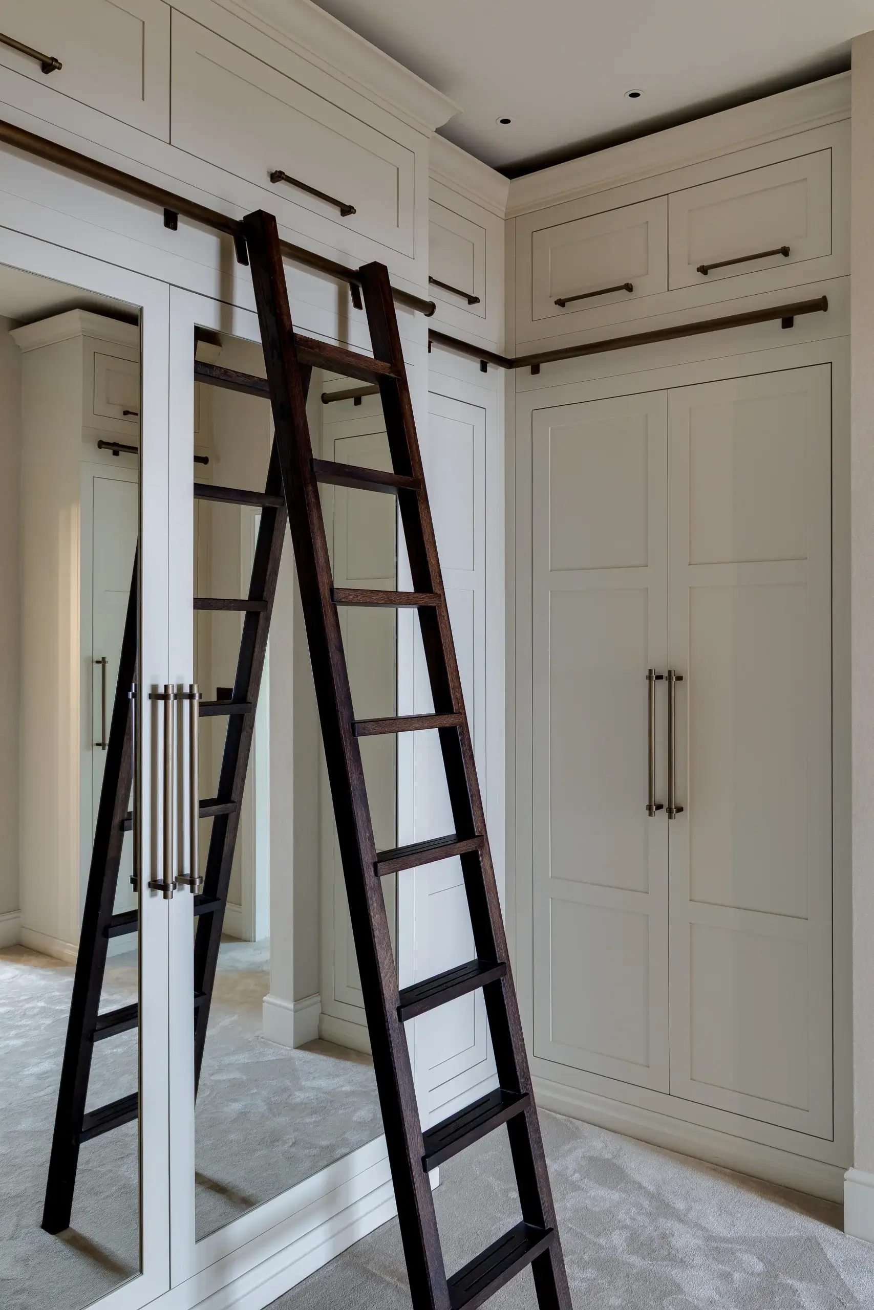 Tall dark wooden library ladder leaning against cream built-in wardrobes with mirrored doors and brass handles.