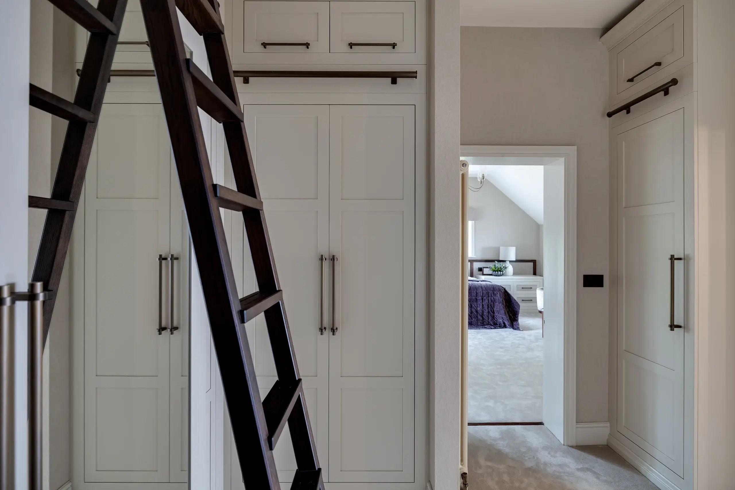 Built-in white closets with long metal handles, a dark wooden rolling ladder, and a view into a bedroom with a purple bedspread.