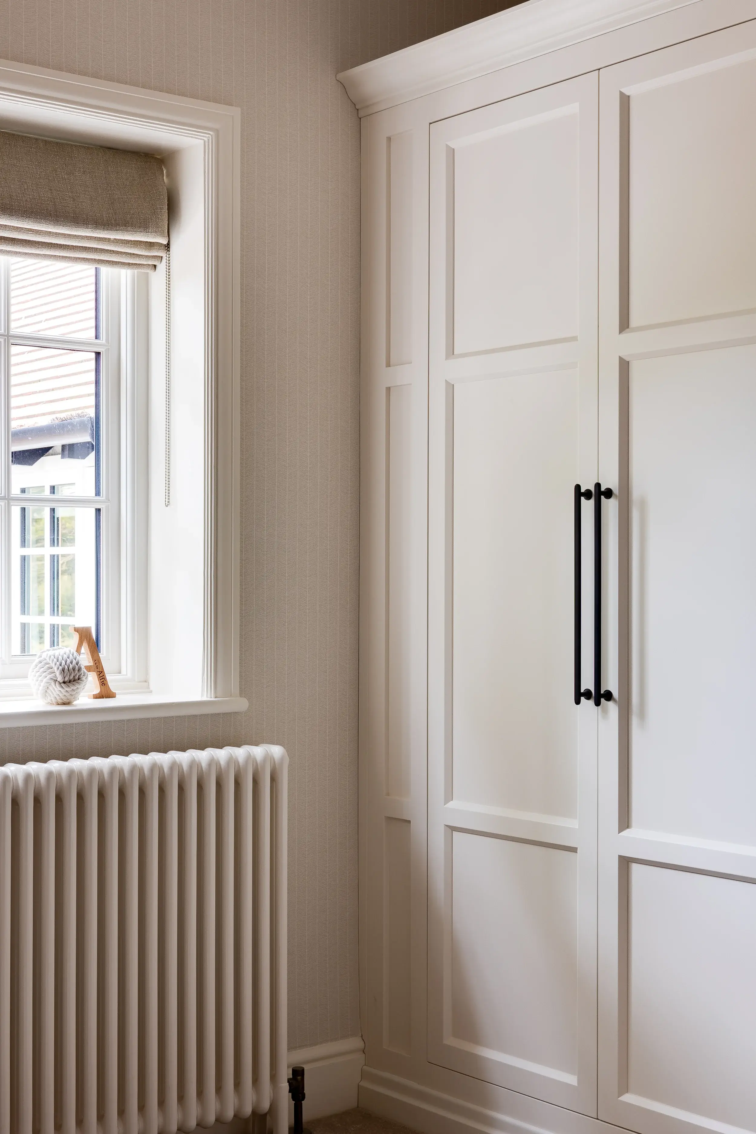 Simple panelled wardrobe in soft cream, with black metal handles, placed beside a sash window and traditional radiator.