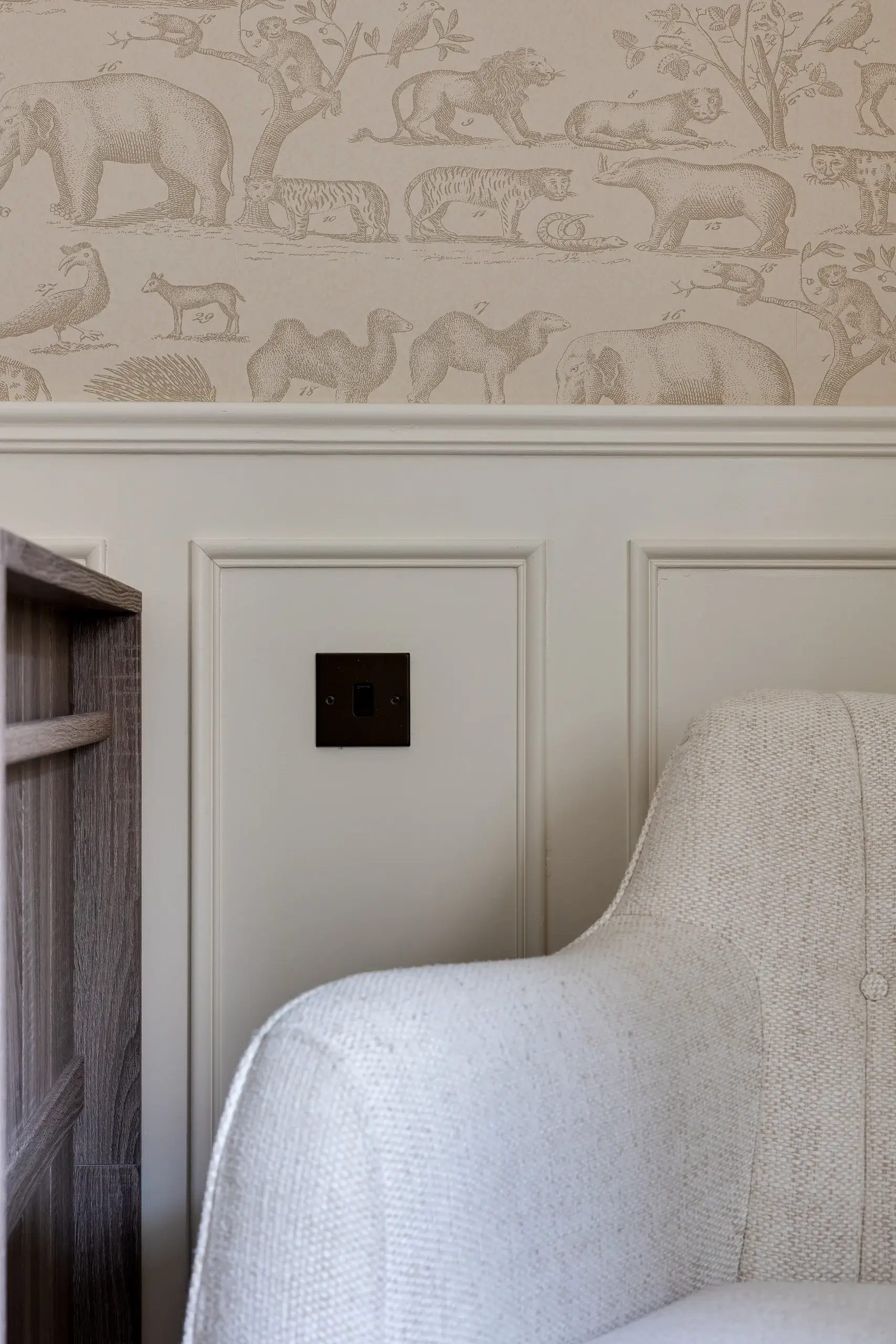 Neutral-toned corner of a room showing white paneled wainscoting, a beige animal-print wallpaper above, a dark square light switch, and a textured off-white armchair arm.