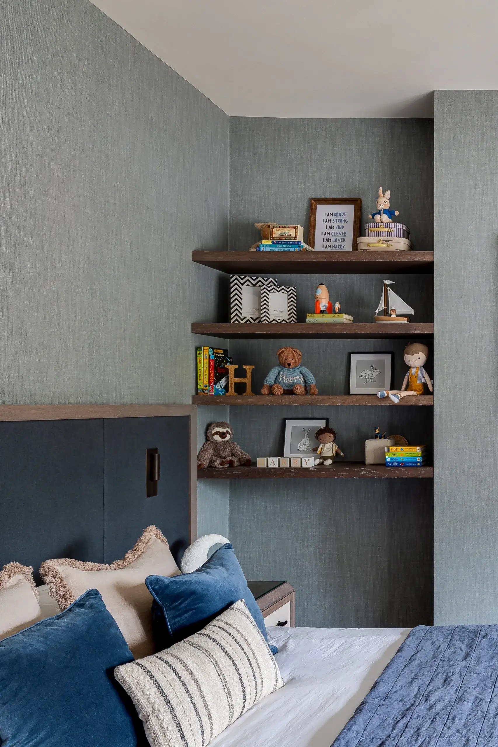 Bedroom corner with built-in dark wood shelves holding children's toys, books, framed prints and a teddy bear above a bed with blue and beige pillows.