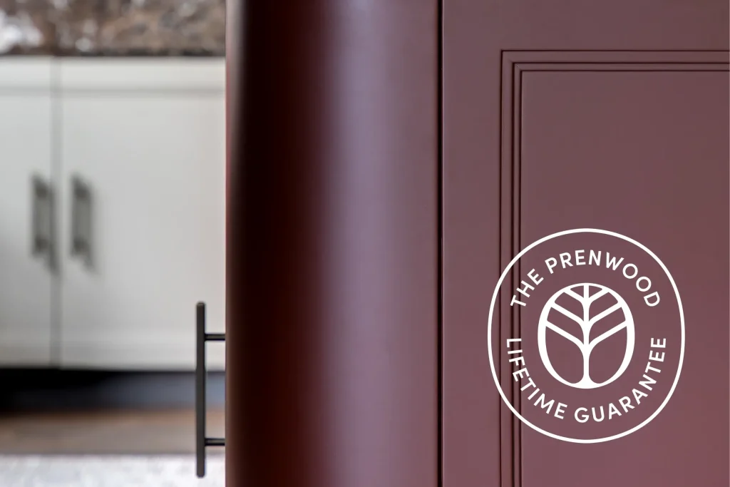 Close-up of a maroon cabinet door with recessed paneling and a vertical black bar handle, white "The Prenwood Lifetime Guarantee" logo on the right, blurred white cabinets in background.