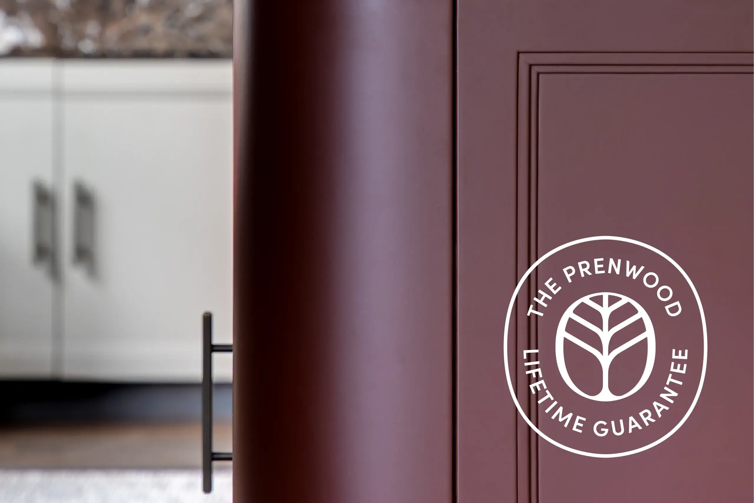 Close-up of a maroon cabinet door with recessed paneling and a vertical black bar handle, white "The Prenwood Lifetime Guarantee" logo on the right, blurred white cabinets in background.