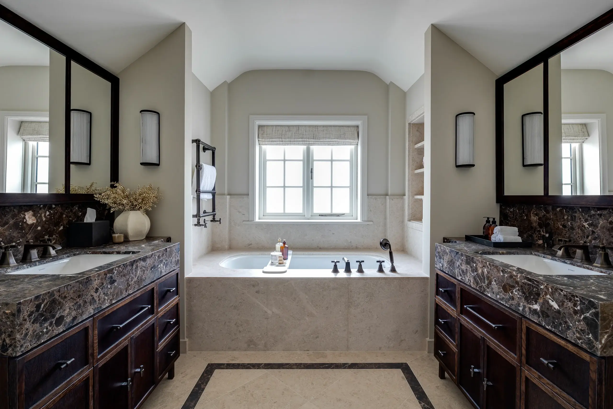 Symmetrical luxury bathroom with central soaking tub under a window, dark marble double vanities and neutral stone finishes