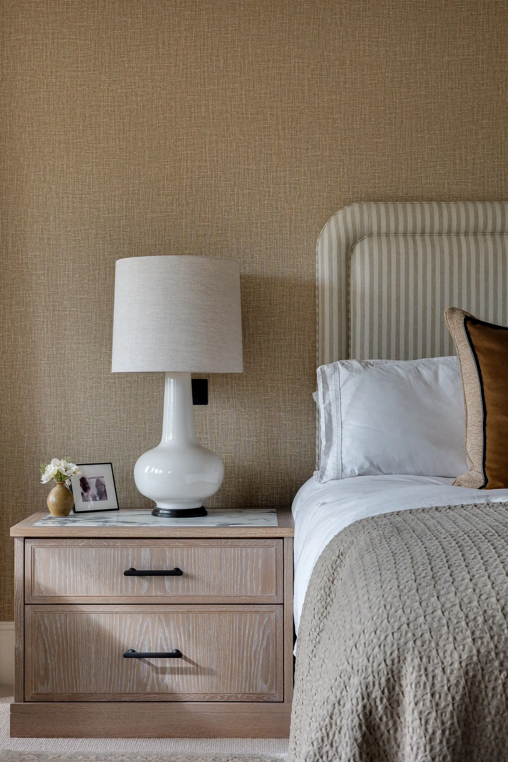 Custom oak bedside cabinet with marble top, paired with a striped upholstered headboard and warm textured wallpaper.