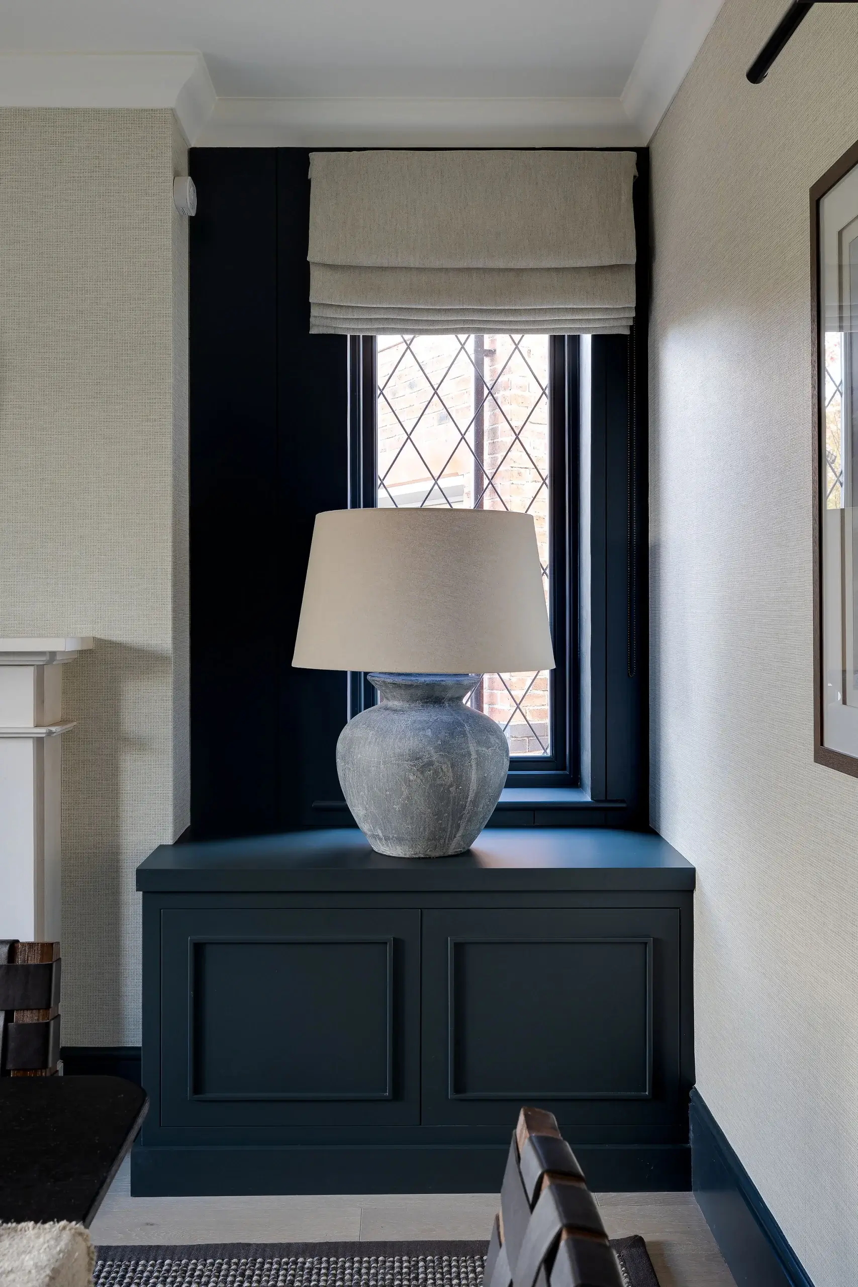 Built-in window seat cabinet in deep blue with panelled joinery, styled with a large stone lamp and Roman blind.