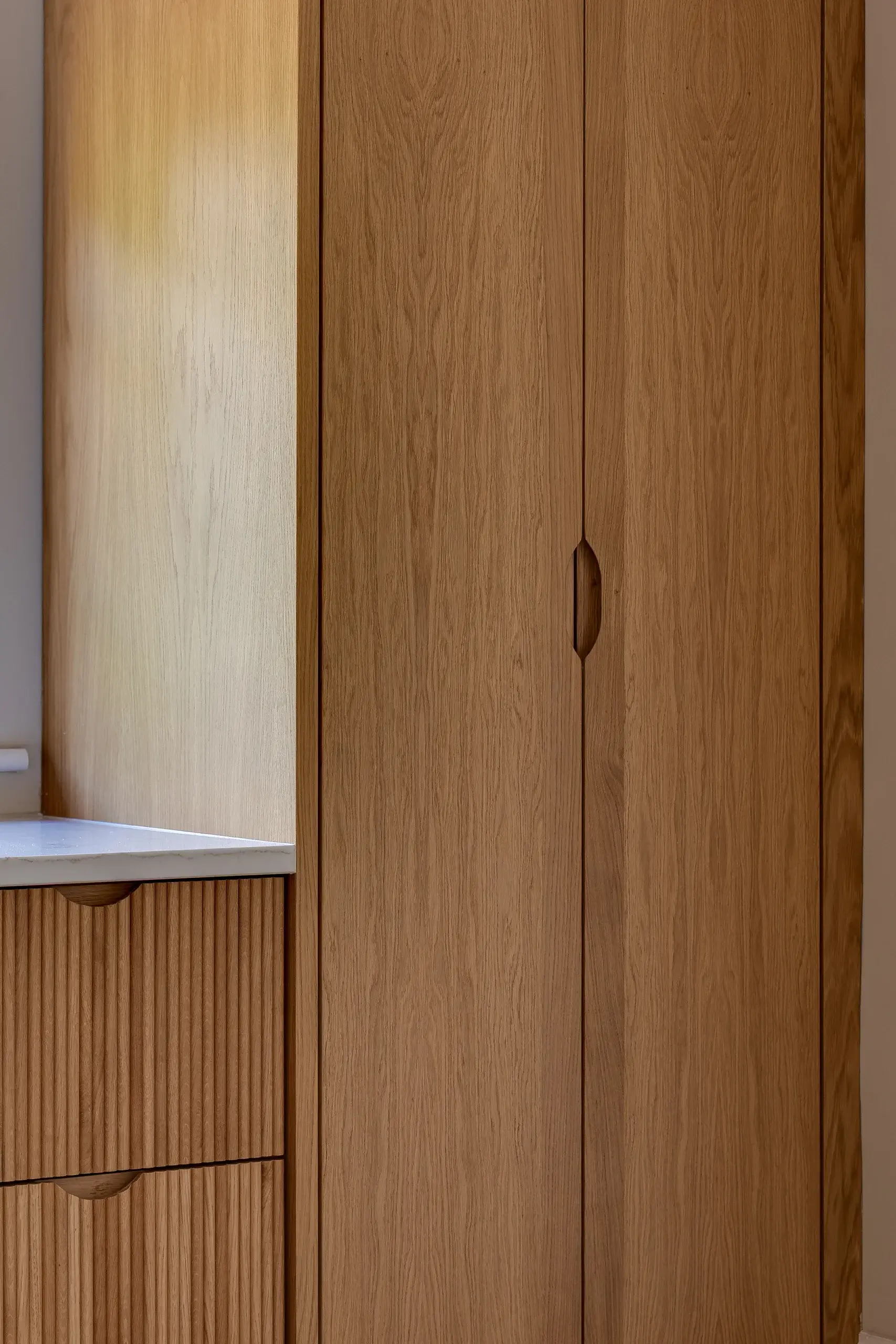 Tall oak cabinet with vertical wood grain and recessed oval pull, next to ribbed wood drawers and a white countertop edge