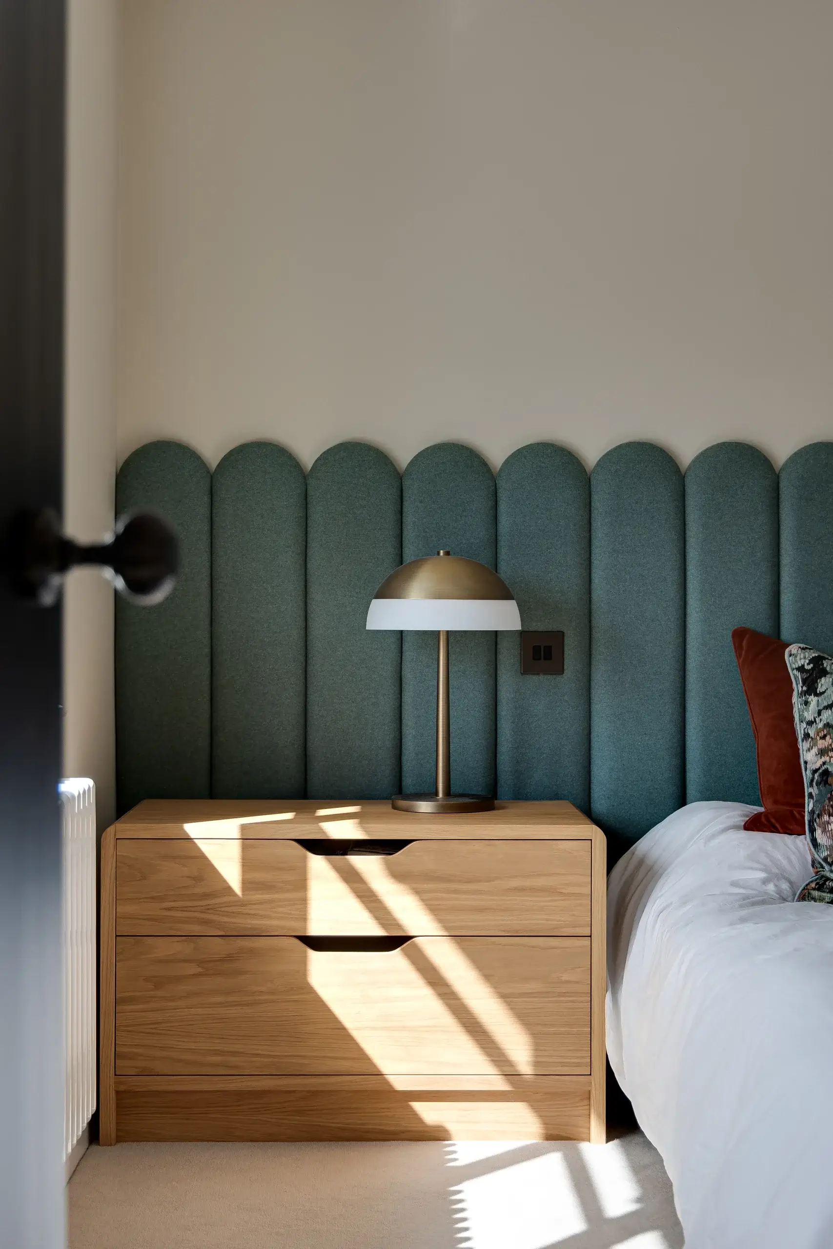 Oak bedside chest with a brass dome lamp in front of scalloped teal upholstered headboard, sunlight on drawers.