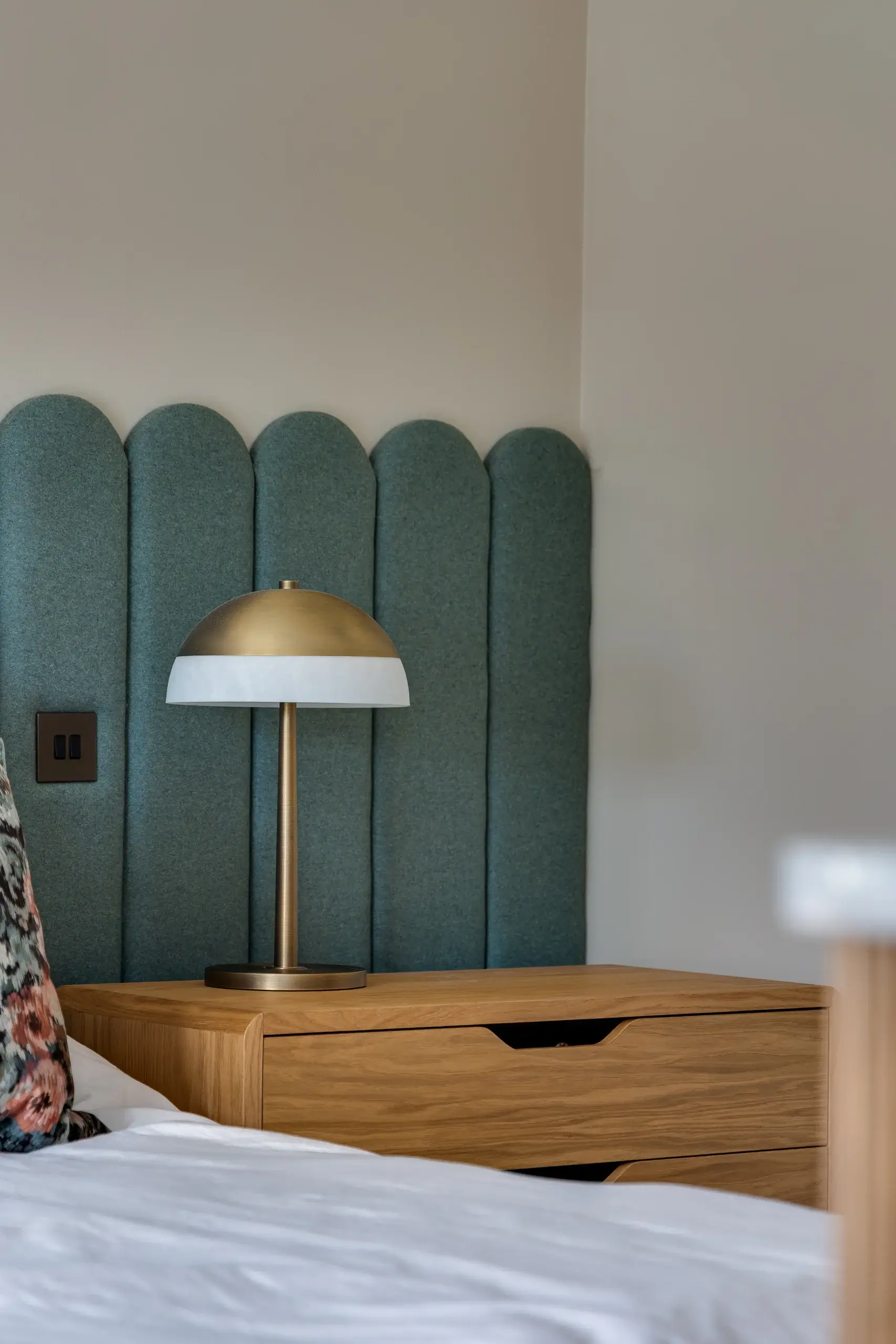 Bedside wooden nightstand with a brass and white dome lamp against a scalloped teal upholstered headboard.