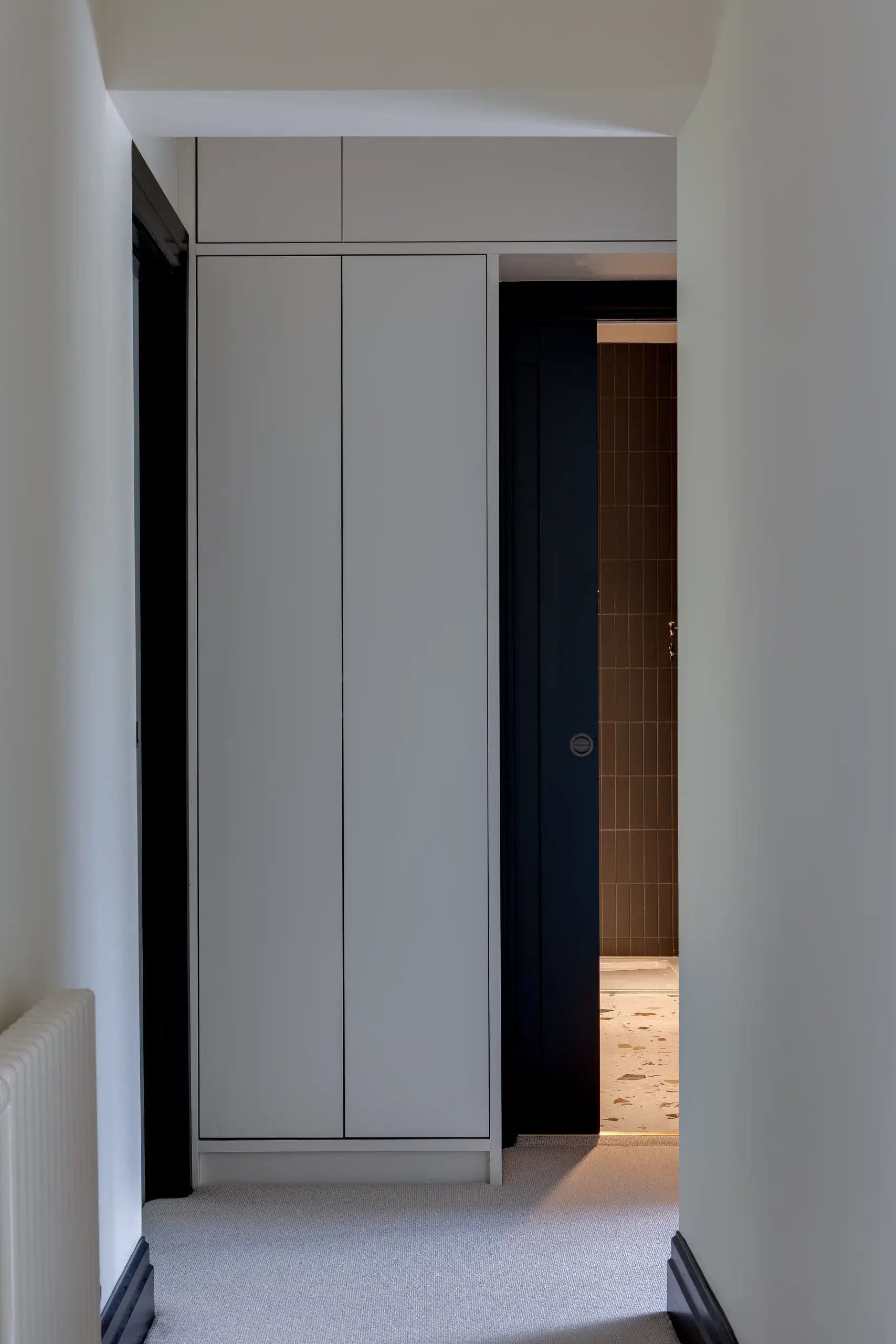 Narrow hallway with pale built-in wardrobe and a dark sliding door partly open to a tiled room with warm floor lighting.