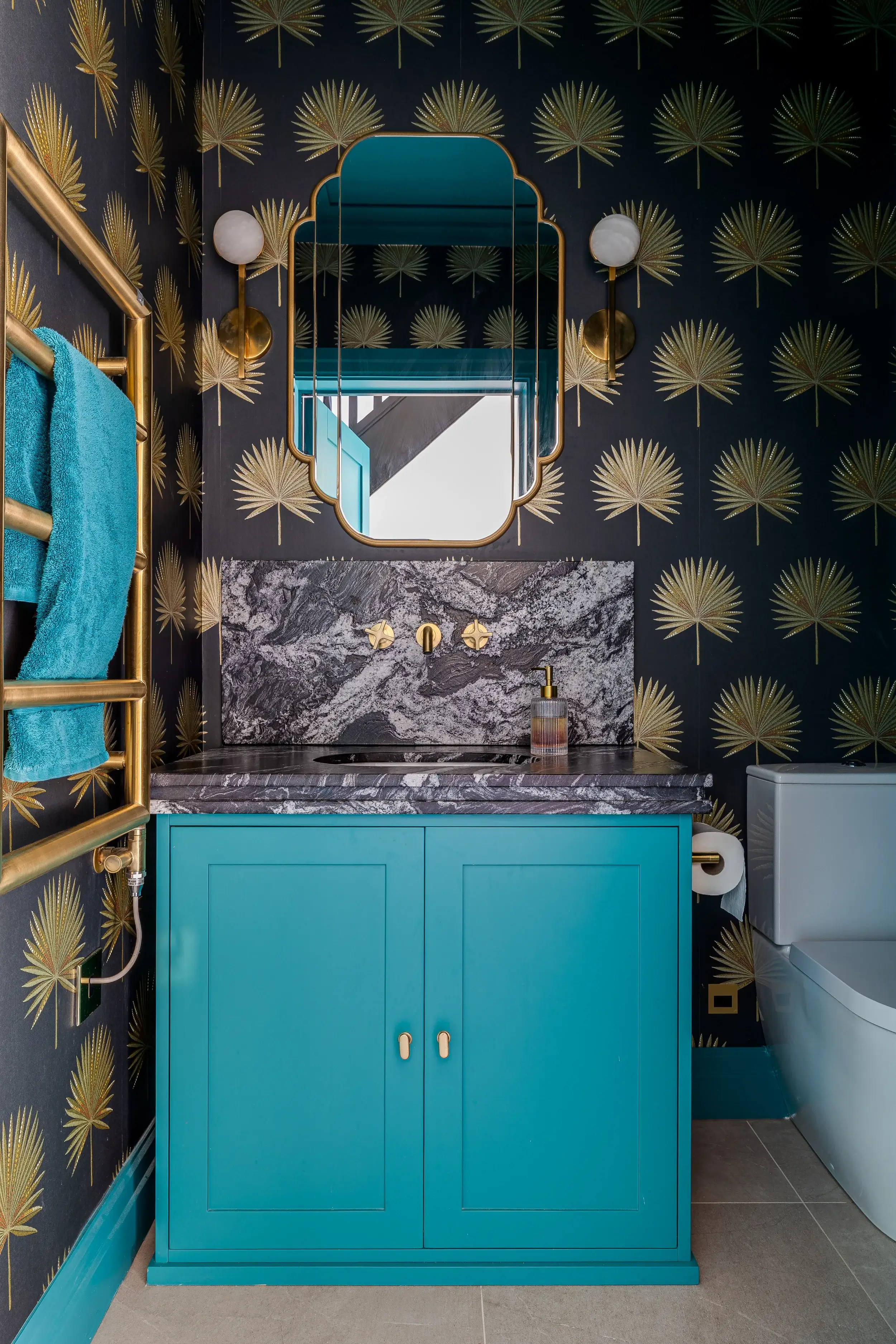 Bold teal vanity, gold palm wallpaper, marble splashback, and art deco mirror framed by brass wall lights.
