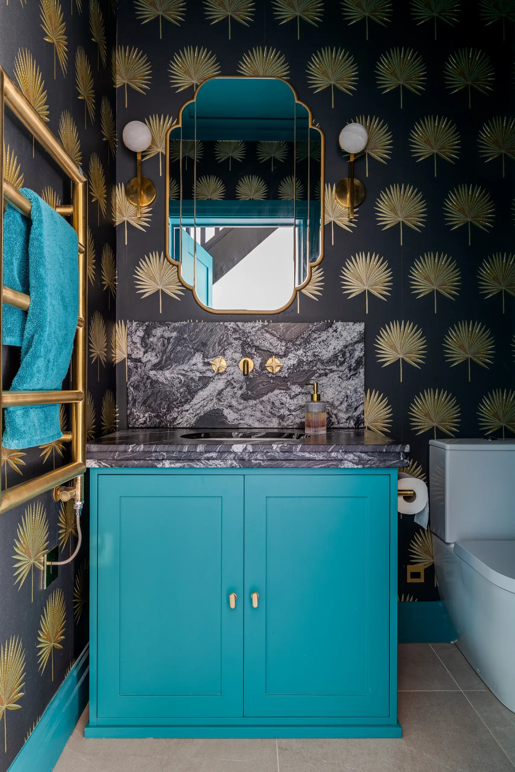 Small teal bathroom vanity with gold hardware, black marble countertop and matching backsplash, gold wall sconces and palm-print black wallpaper.