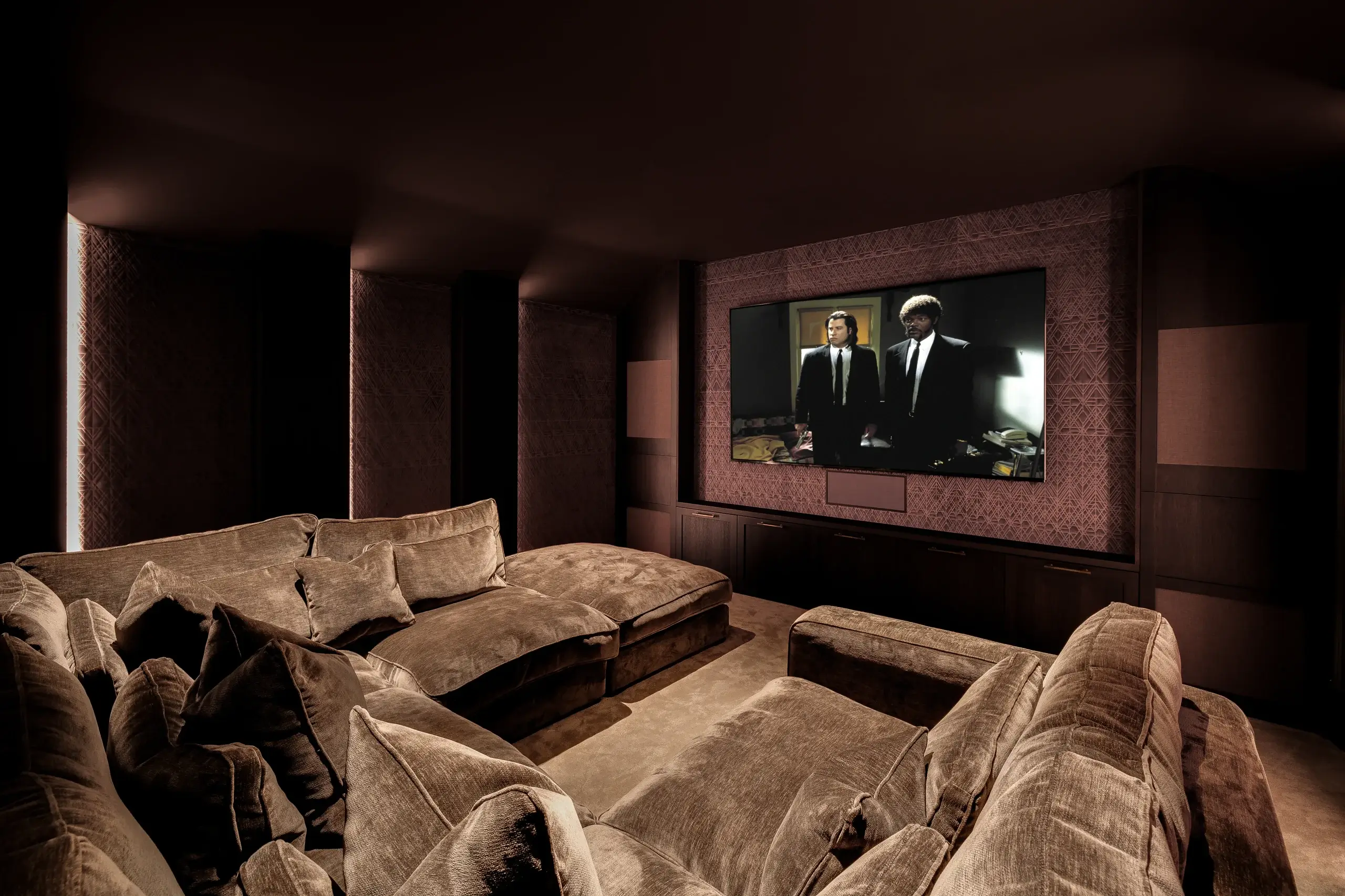 Plush, dimly lit home theater with oversized brown velvet sectional facing a large wall-mounted TV showing two men in suits.