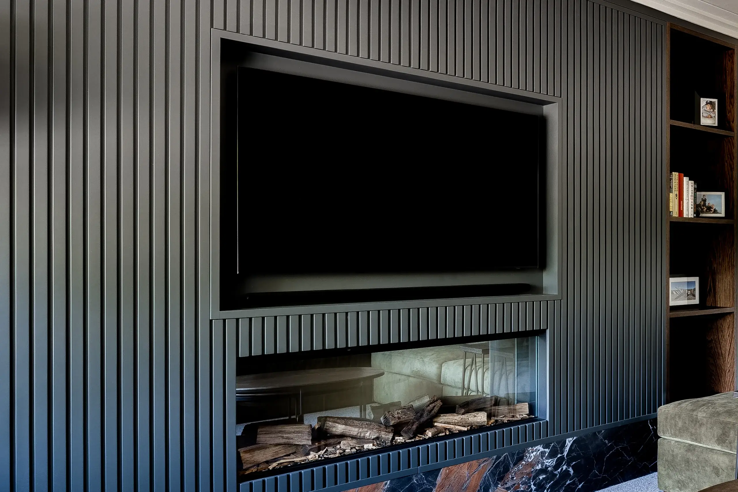 Built-in dark gray paneled wall with a recessed flat-screen TV above a modern glass-front gas fireplace and wood log display, adjacent to wooden shelving.