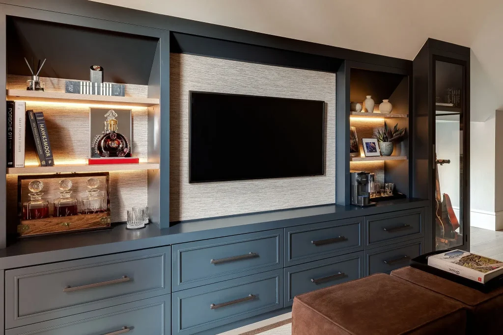 Built-in dark blue media cabinet with a wall-mounted flat-screen TV, lit display shelves holding books, decanters, plants and a coffee station, and lower drawers.