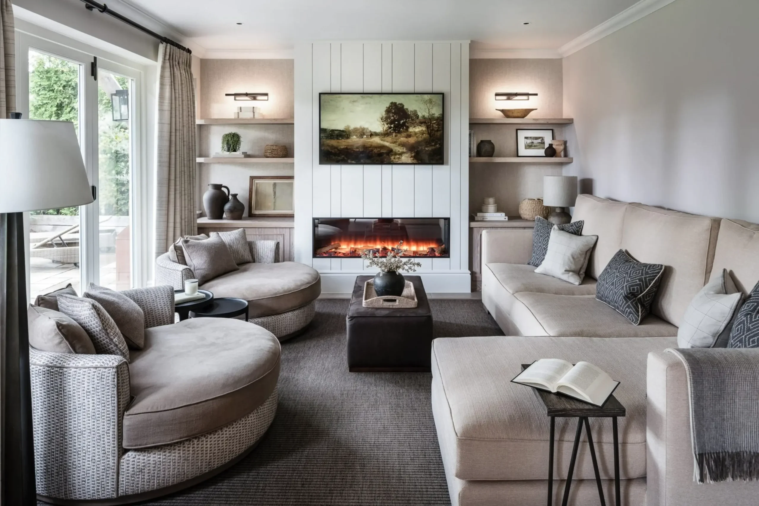 Cozy neutral-toned living room with curved lounge chairs, large sectional sofa, wall-mounted electric fireplace under a landscape painting, and built-in shelving.