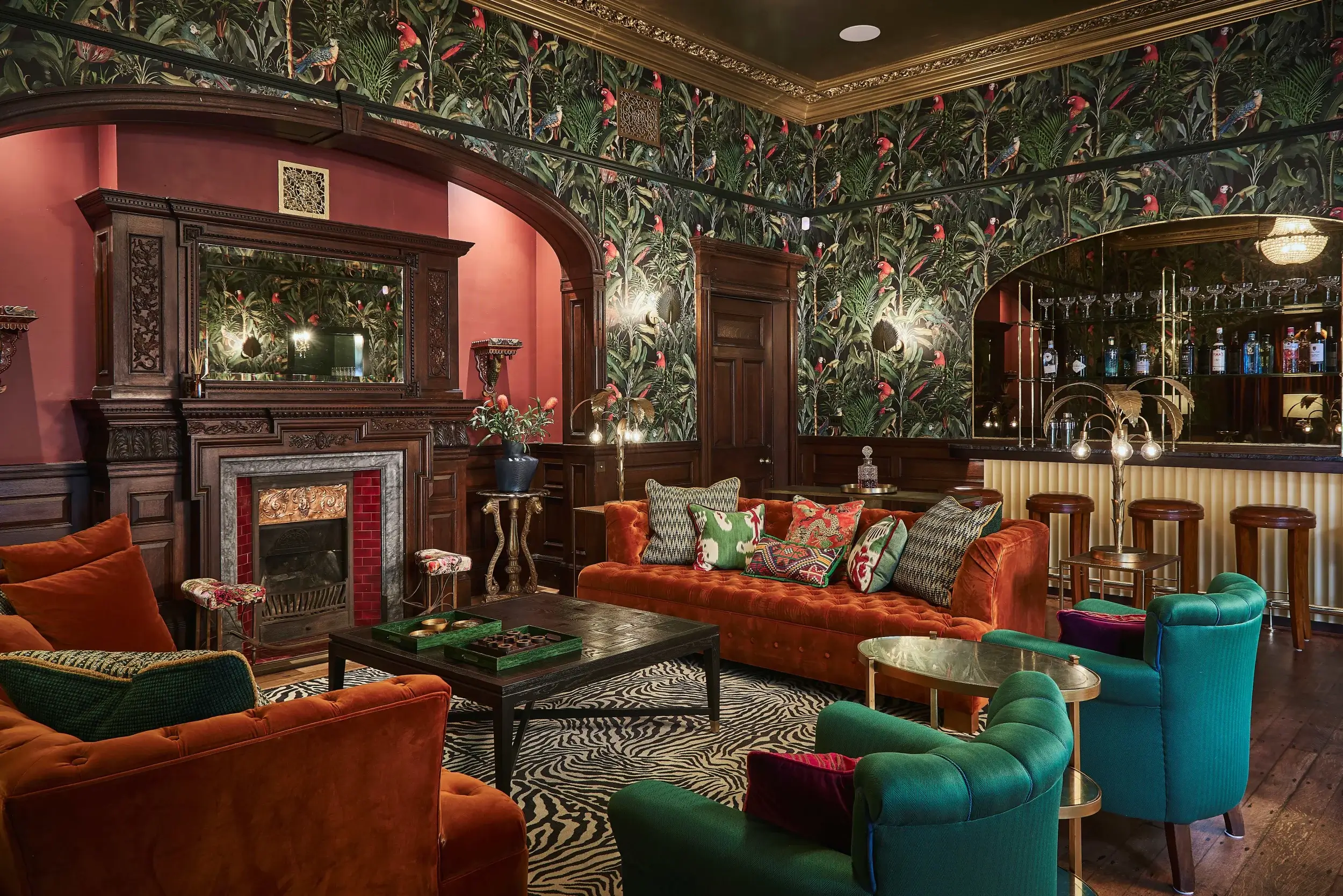 Richly decorated lounge featuring an ornate carved wood fireplace, orange velvet sofa, patterned cushions, and a mirrored bar area in the background.