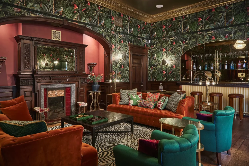 Victorian-style lounge with velvet orange sofas, teal armchairs, patterned wallpaper with parrots, ornate fireplace, and bar.