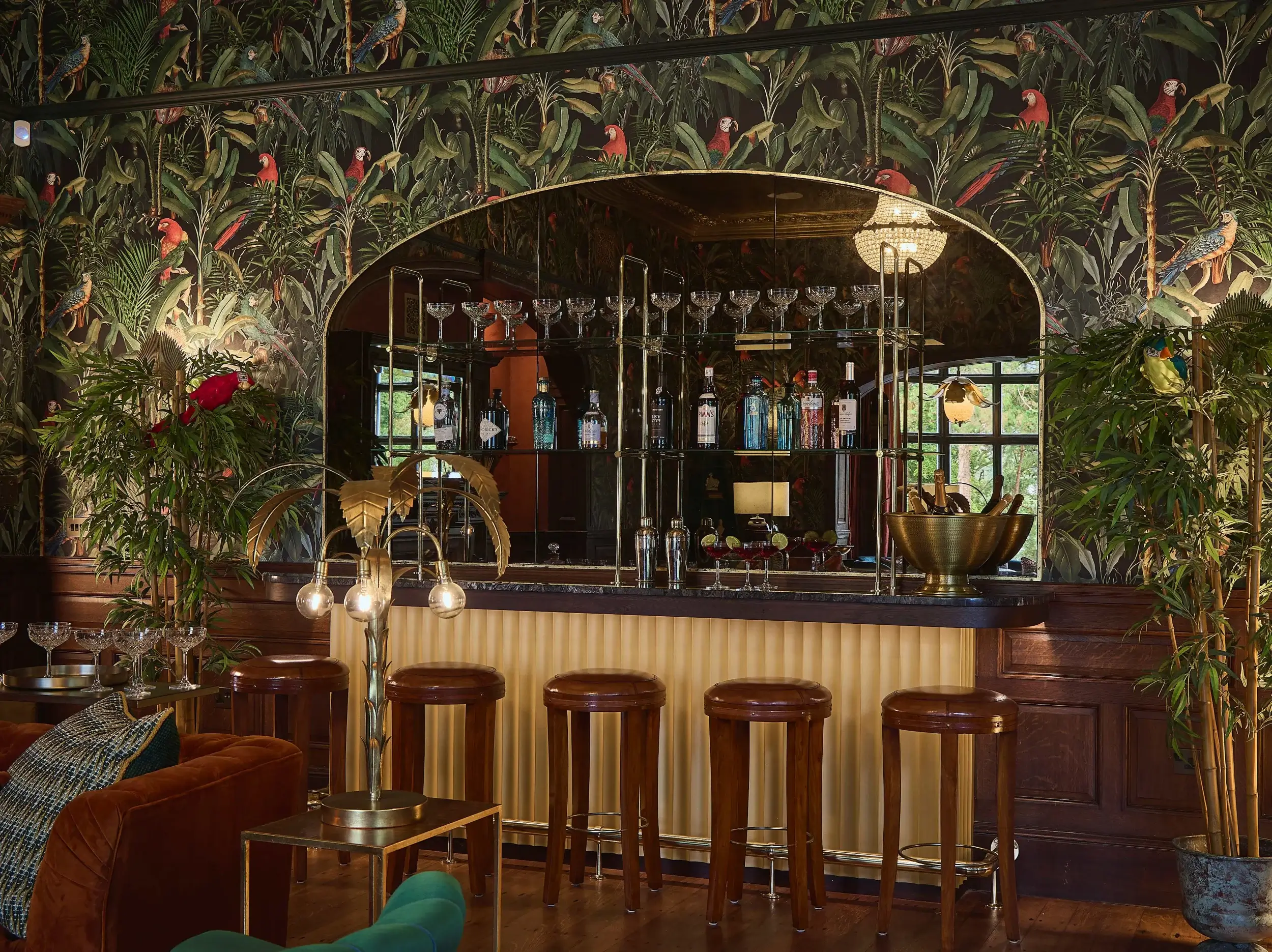 Elegant bar area with mirrored shelving, wooden bar stools, gold accents, and tropical bird wallpaper surrounding the space.