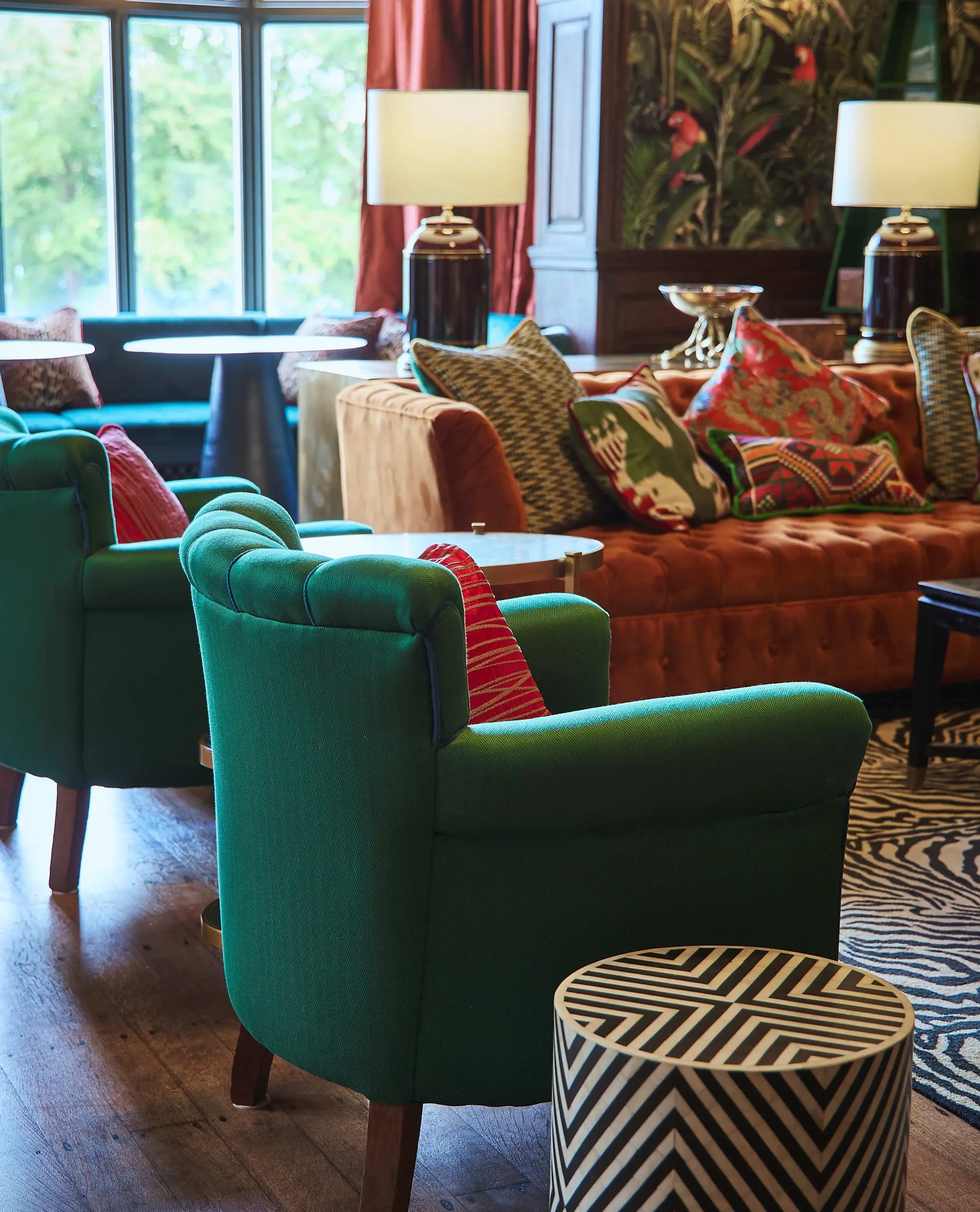Detail of the lounge seating with emerald green armchairs, patterned throw pillows, and a zebra-print rug beside the orange velvet sofa.