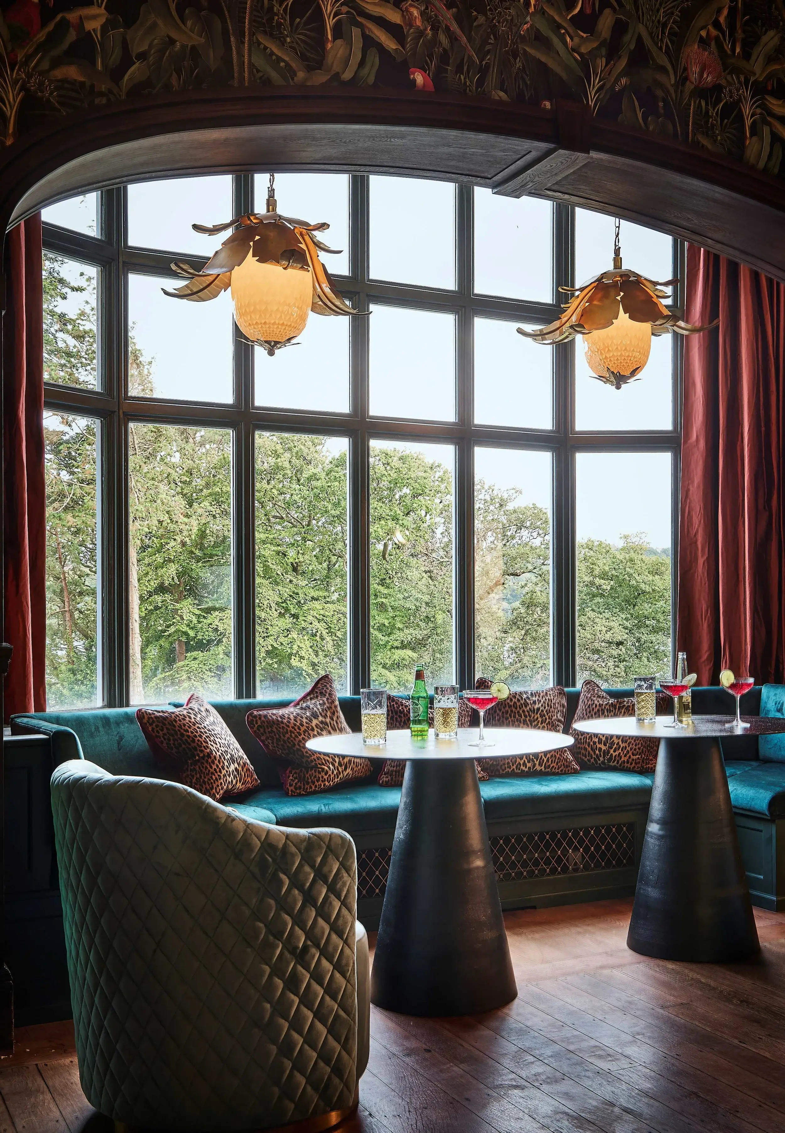 Cozy window seating nook with teal banquette seating, round black tables, leopard-print cushions, and floral-inspired pendant lighting.
