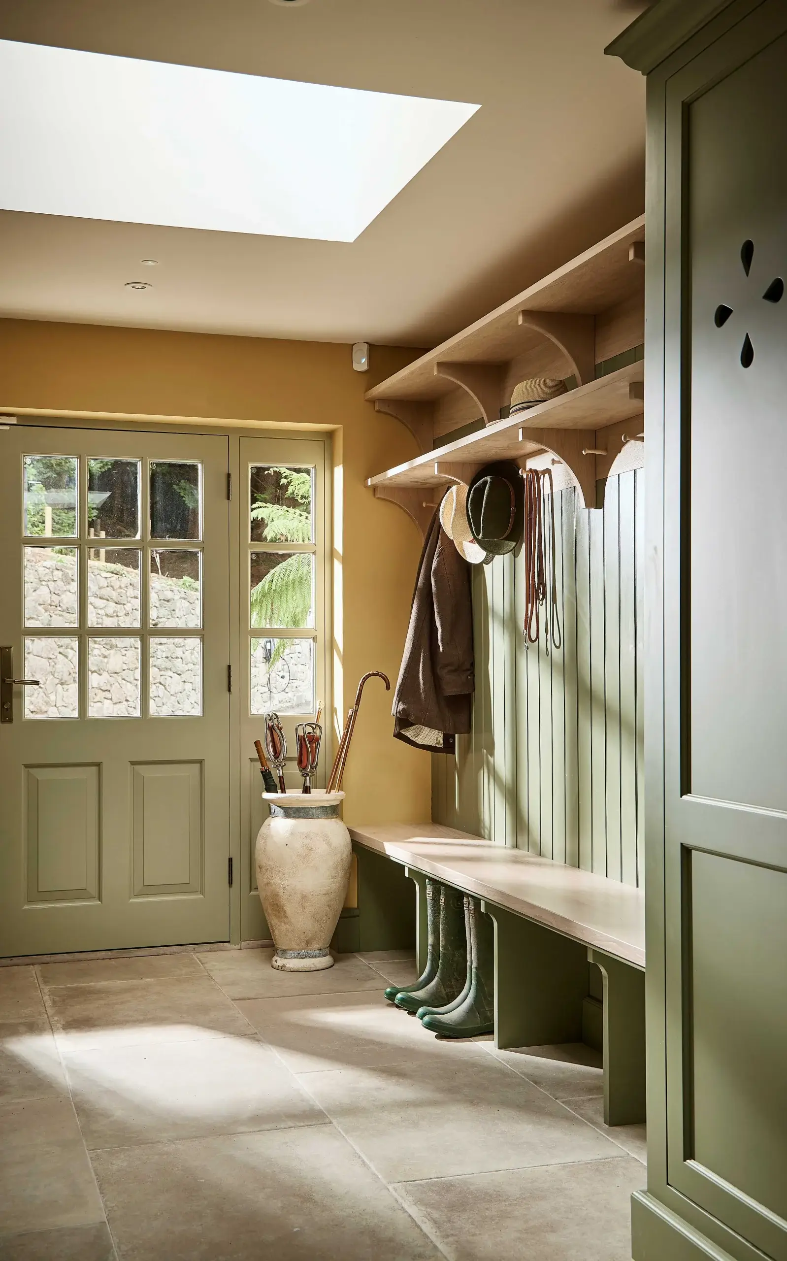 Mudroom with sage-green built-in bench and hooks, tall vase holding umbrellas, boots tucked under bench, glass-paneled door and skylight