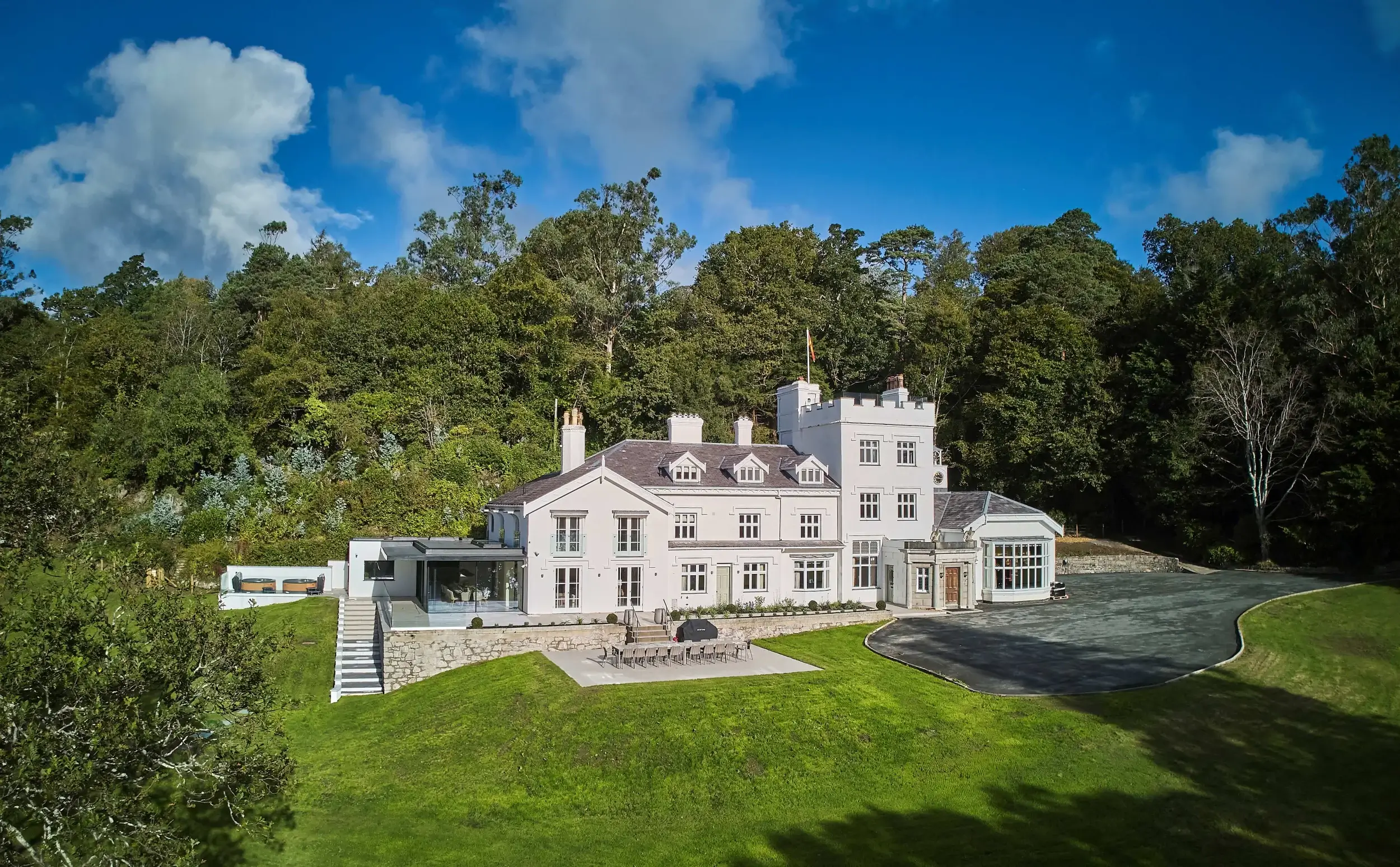 Elegant white manor house with an outdoor terrace and stone retaining wall, framed by lush greenery and blue skies.