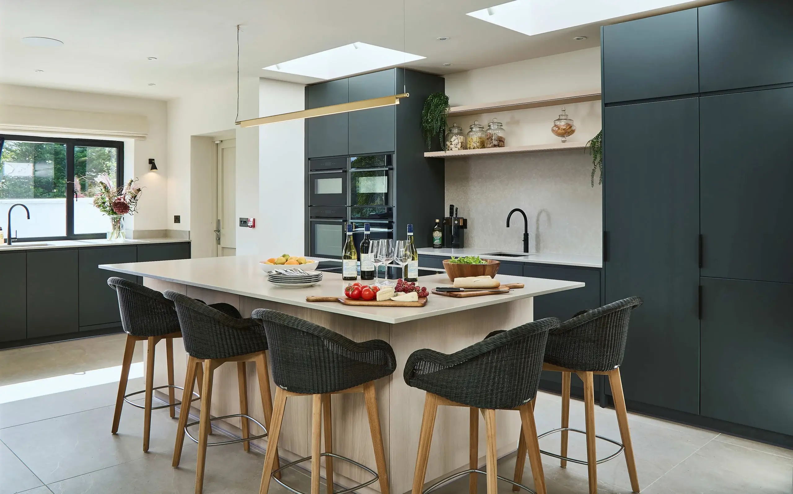 Modern kitchen with a large island, five woven bar stools, dark cabinets, skylights, and food and wine set on the countertop