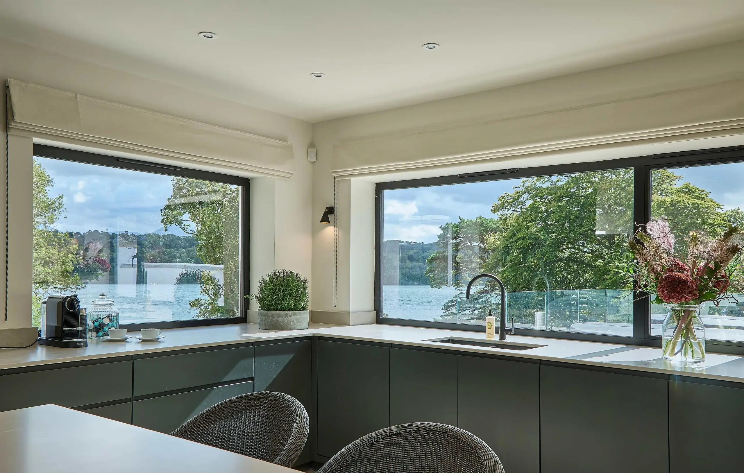 Modern kitchen corner with large windows overlooking a lake, dark cabinets, countertop sink, coffee machine, and vase of flowers