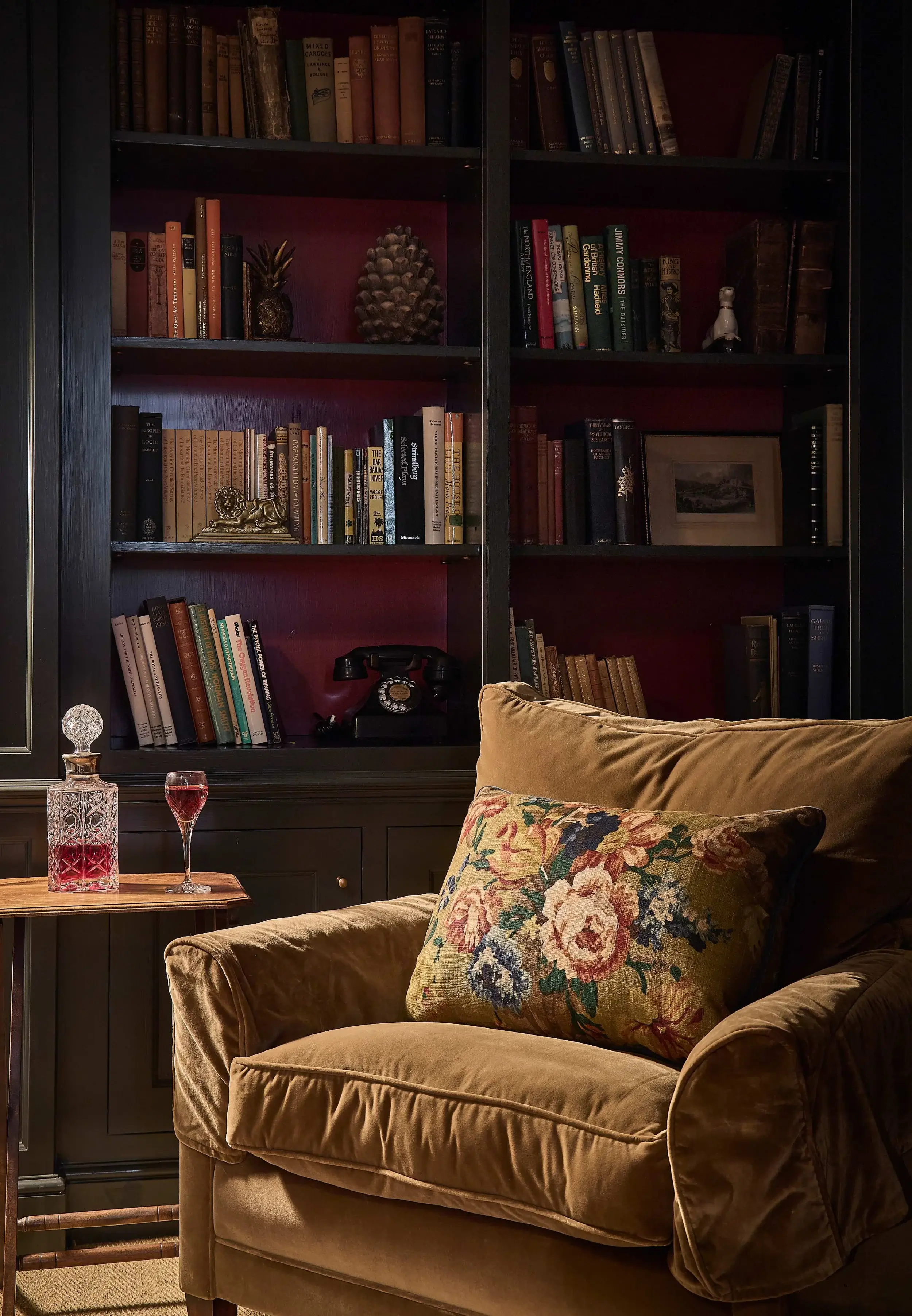 Plush brown armchair with floral cushion beside a small table holding a crystal decanter and glass, in front of a book-filled dark wood bookshelf