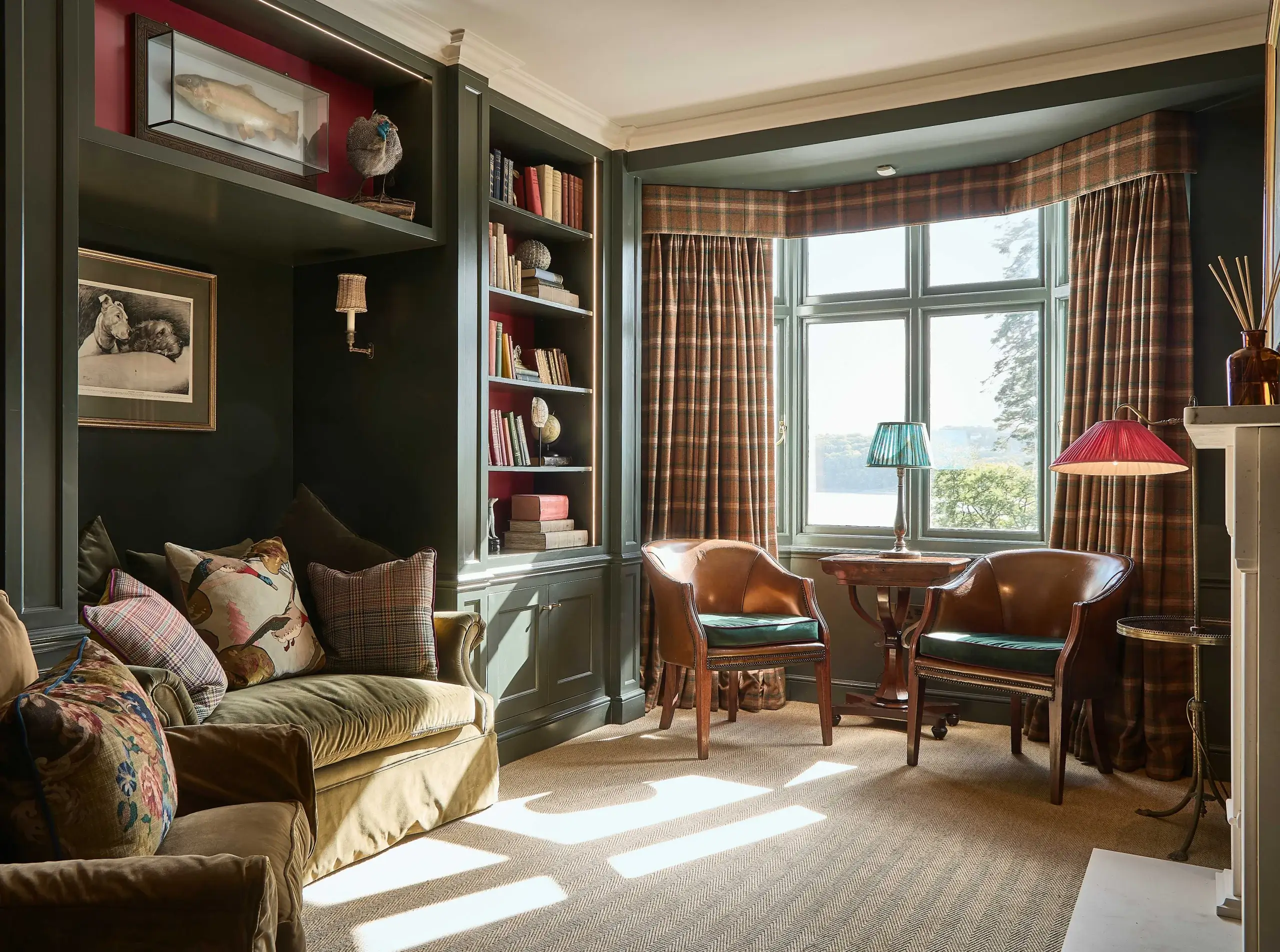 Cozy reading nook with green built-in bookshelves, plaid curtains, velvet sofa with patterned cushions, and two leather armchairs by a sunlit bay window.